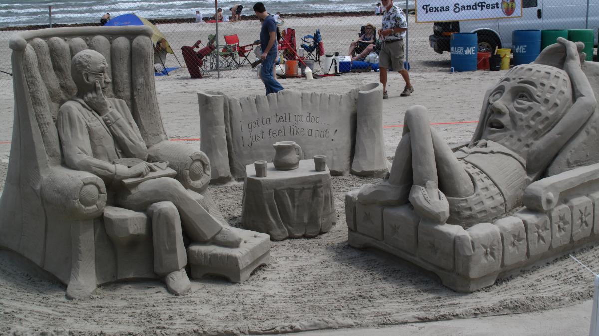 Sand sculpture depicting a psychiatrist sitting in a chair on the left. To his right is a peanut with a face laying on a couch. In between them is a table of sand and a scroll that says, "I gotta tell you, Doc. I just feel like a nut!"