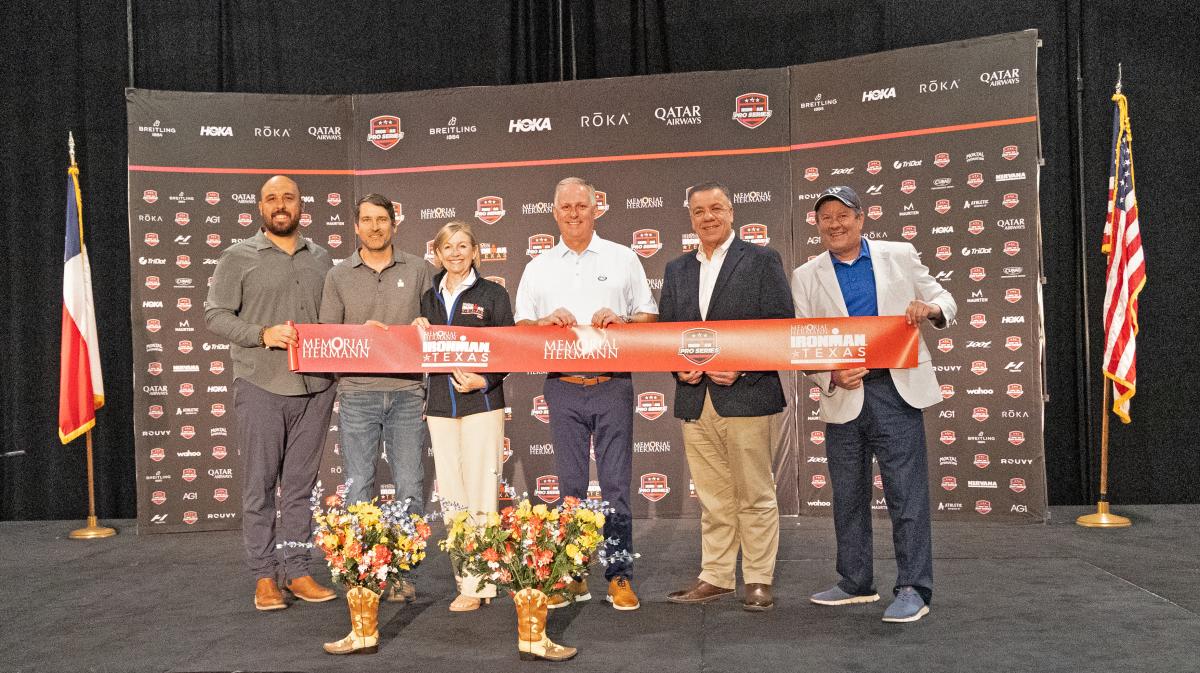 Six people on a stage hold a thick red ribbon that reads, "MEMORIAL HERMANN," "MEMORIAL HERMANN IRONMAN TEXAS," and "IRONMAN PRO SERIES" in repeat across it. In the middle are Linda Nelson and Brad Bailey. On the far right is Nick Wolda.