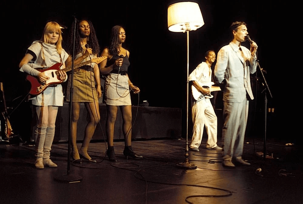 A band performs on stage under warm lighting. At the front, a man in a light-colored suit sings into a microphone. To his left, three women stand at microphones, one playing an electric guitar while the others appear to be backing vocalists. Another musician plays guitar in the background. A tall floor lamp stands on stage among them, adding a unique, living-room-like element to the setup.