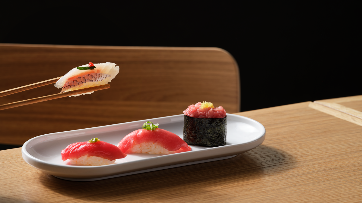 Close-up image of two chopsticks holding a sushi roll with three other pieces of sushi sitting on a plate.
