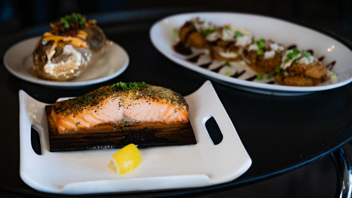 An enticing photo of a serving tray featuring salmon with a wedge of lemon, a baked potato, and fried green tomatoes drizzled with balsamic reduction plated beautifully on white plates of various sizes and shapes.