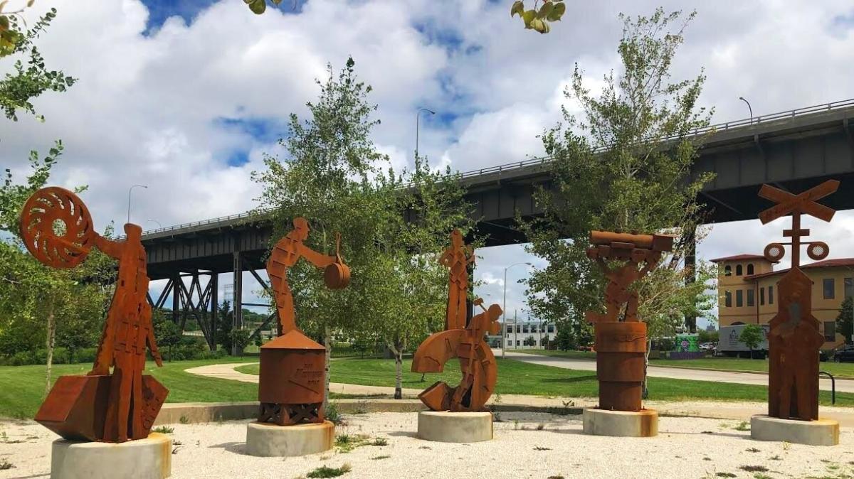 Six rust-colored steel sculptures stand in a semicircle on a gravel pad beneath a tall overpass in Milwaukee’s Menomonee Valley. Each figure represents a different role in railroad history, including workers with tools and symbols like wheels, drums, and crossing signs. Trees and grassy paths surround the installation, titled People of the Road, with a mix of industrial and natural elements in the background.
