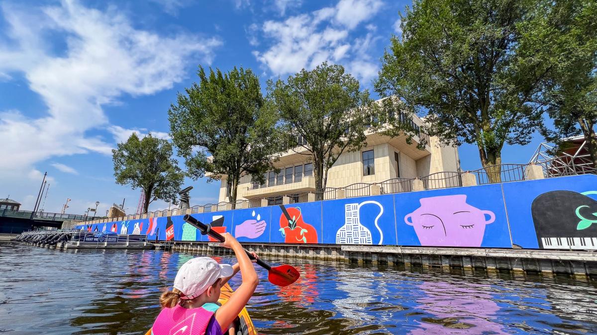 Child paddling a kayak on a sunny day along a river with colorful mural art and trees lining the waterfront.