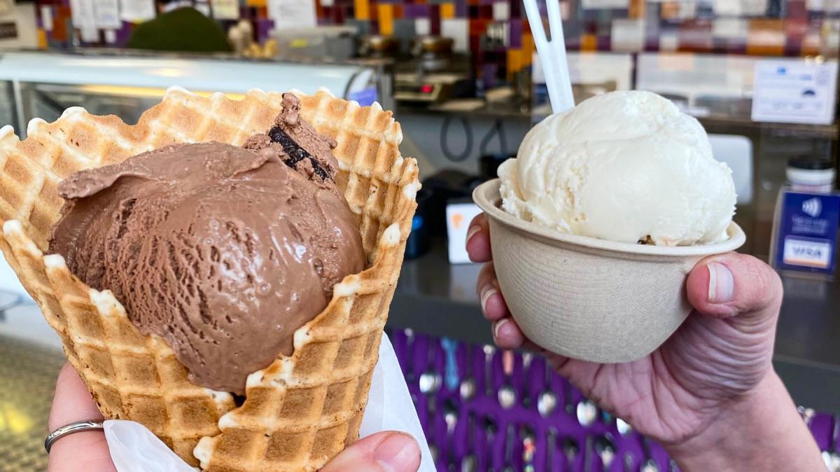 Close-up of two hands holding ice cream: one with a waffle cone filled with chocolate ice cream and another with a cup of vanilla ice cream.