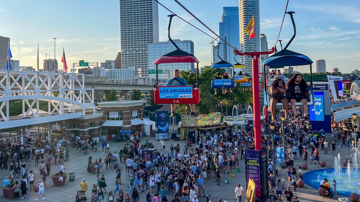 Crowds enjoying a lively outdoor festival with food stands and a sky ride, with people seated on chairlifts overlooking the scene and city skyline.