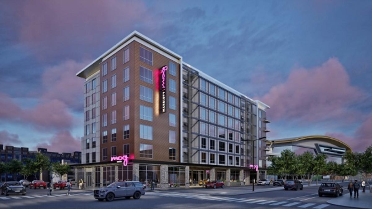A rendering of the upcoming Moxy Hotel in Milwaukee’s Deer District, featuring a modern multi-story building with floor-to-ceiling windows and vibrant pink signage. The hotel is surrounded by street traffic and pedestrians, with the Fiserv Forum visible in the background under a colorful evening sky.