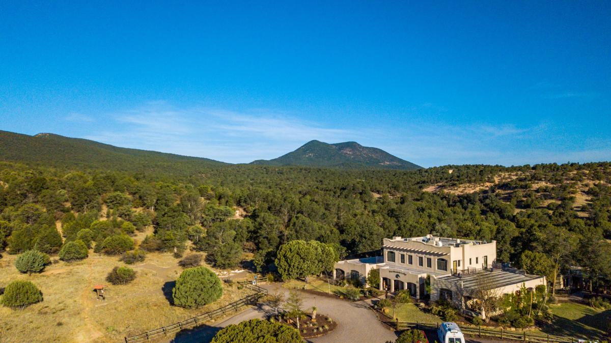 Aerial view of a large estate with a two-story building, surrounded by expansive green fields and a dense forest, with a mountain in the background under a clear blue sky.