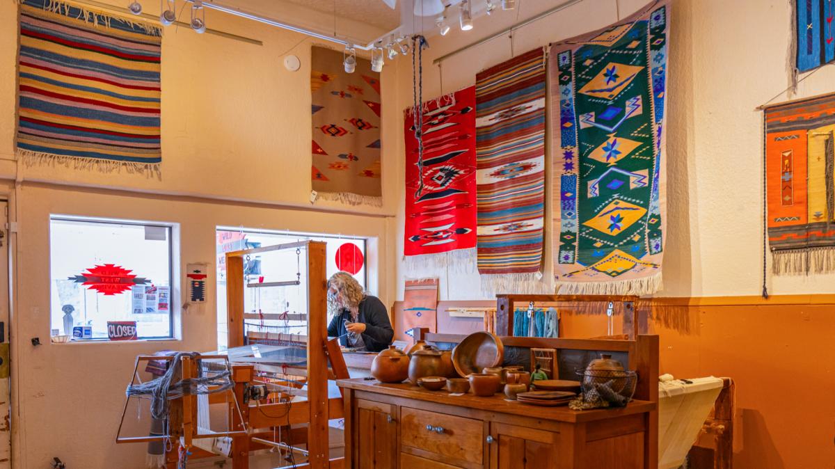 a woman stands behind a wooden loom, a wooden dresser is in the fore ground and there are weaved rugs adorning the walls