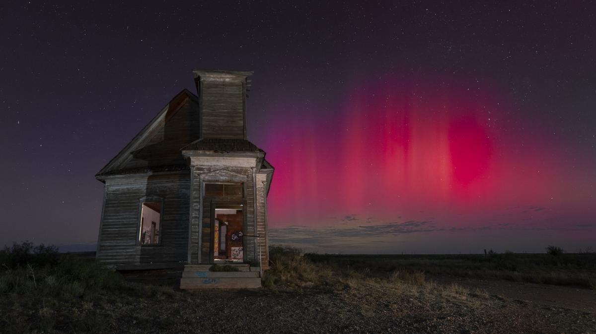 A weathered, abandoned house stands in front of a vivid red aurora under a starry sky.