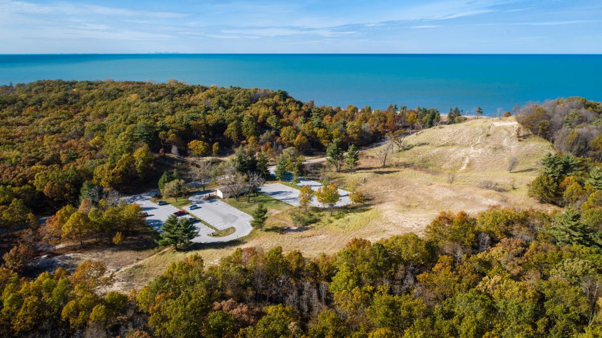 Aerial perspective of a lakeside nature area with a small parking lot, golden dunes with visible trails, and a dense forest with autumn leaves. The vast lake with a blue hue extends to the horizon beneath a partly cloudy sky.
