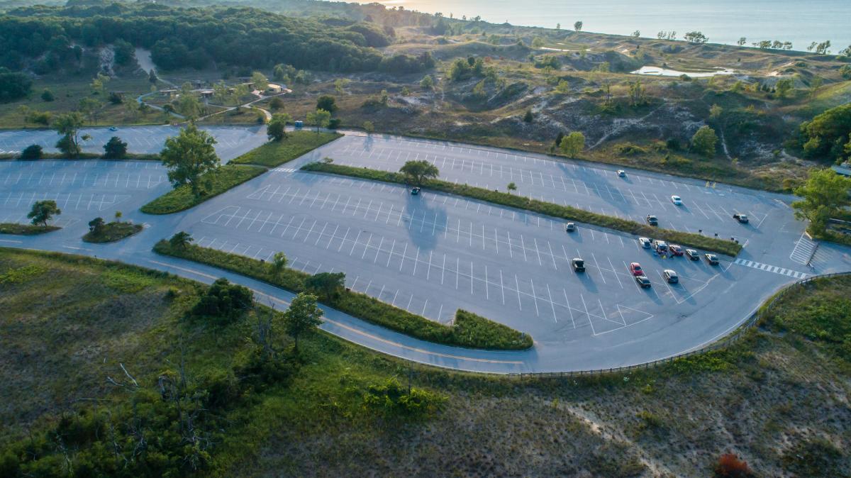 Aerial view of a large parking lot surrounded by dunes and vegetation. Lake Michigan is seen in the background.