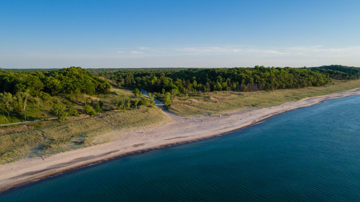 Aerial view of the shoreline. The water is a deep blue. The sand changes to beach grass and eventually trees.