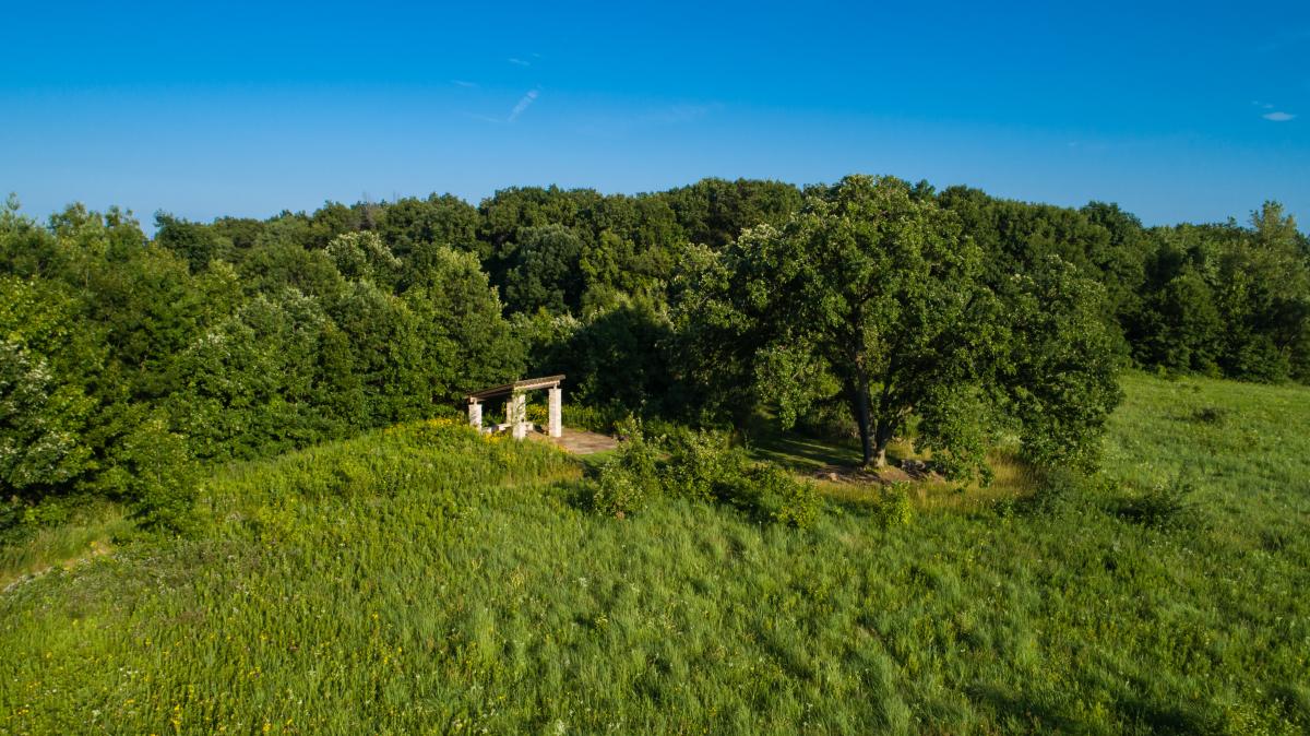 An aerial view of a green landscape and bright blue sky. A stone structure is in the center of the photo.