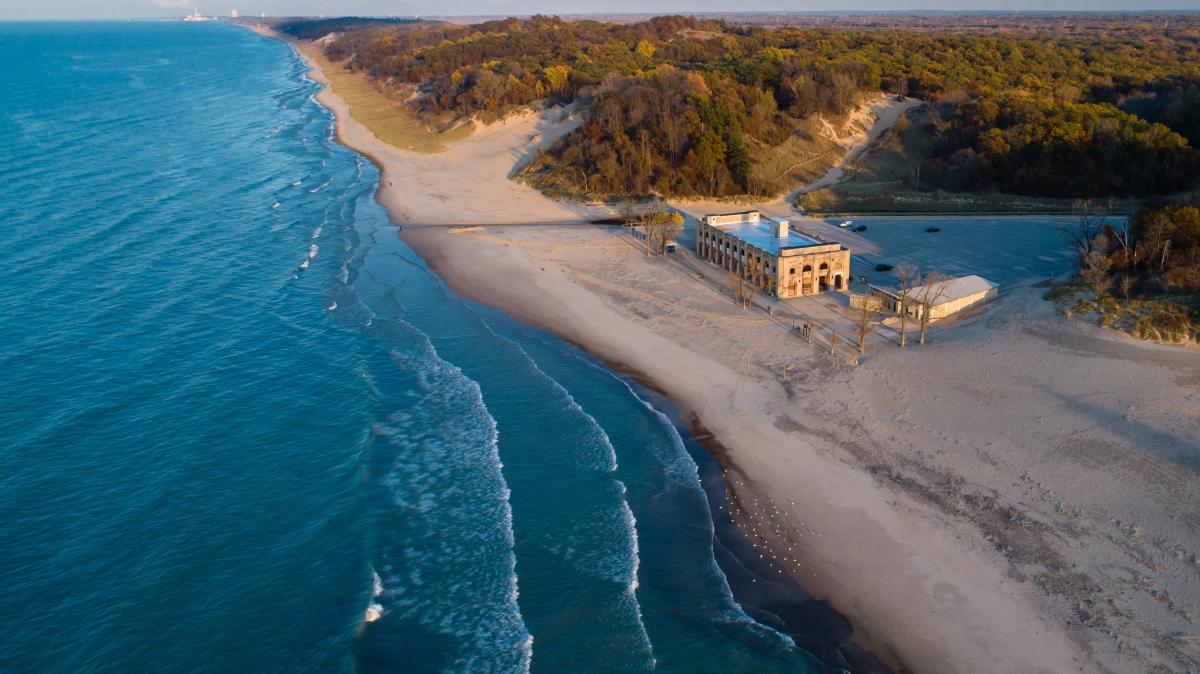 Bright blue water with white cresting waves near the shore sits to the left. On the right is a long sandy beach that spreads to the horizon line. Wooded dunes extend from the beach and into the distance. A stone building sits on the beach with a parking lot behind it.