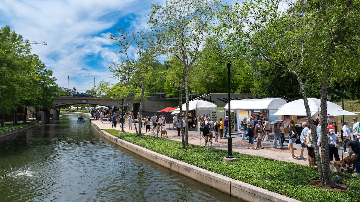 White tents and umbrellas line The Woodlands Waterway to shade displays of local and international artwork for sale at The Woodlands Waterway Arts Festival. Dozens of people of all ages take in the sights.