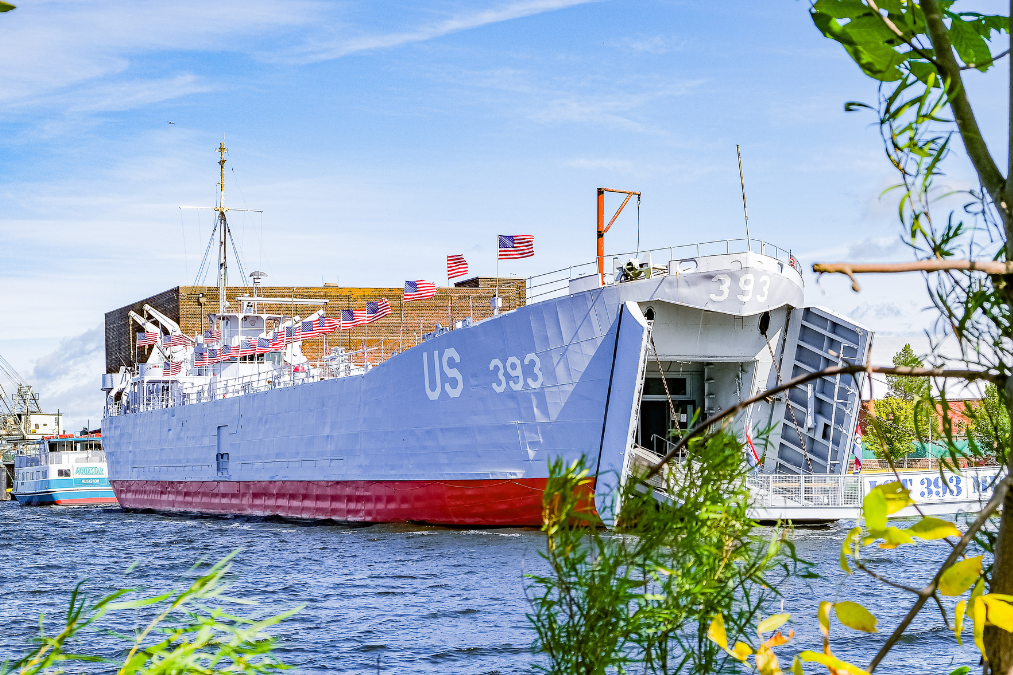 Historic US 393 naval ship docked on blue water, decorated with American flags, viewed through leafy branches under a bright sky.