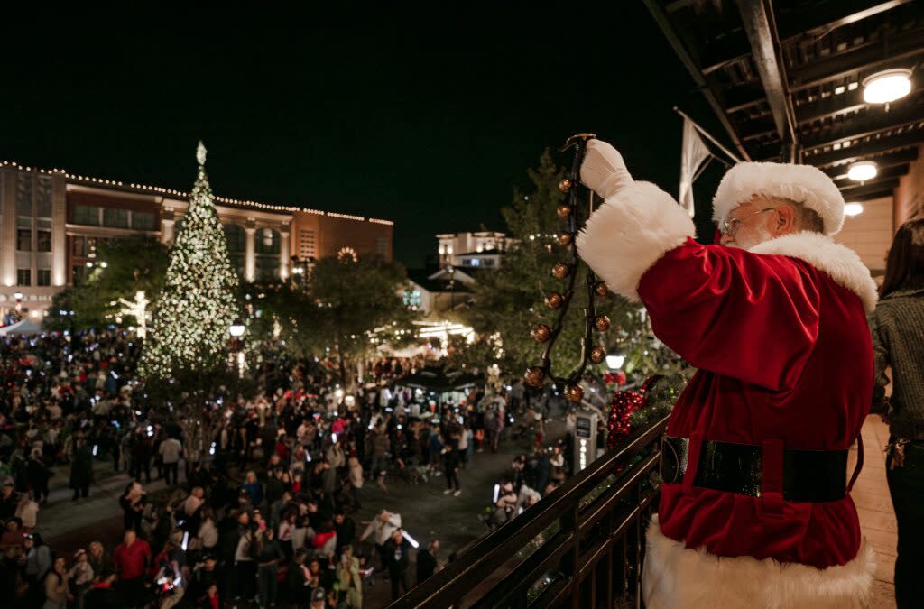 The viewer stands to the left of Santa, who waves jingle bells to a crowd from the roof of a building at Market Street, which is lit up with Christmas lights and holiday activities.