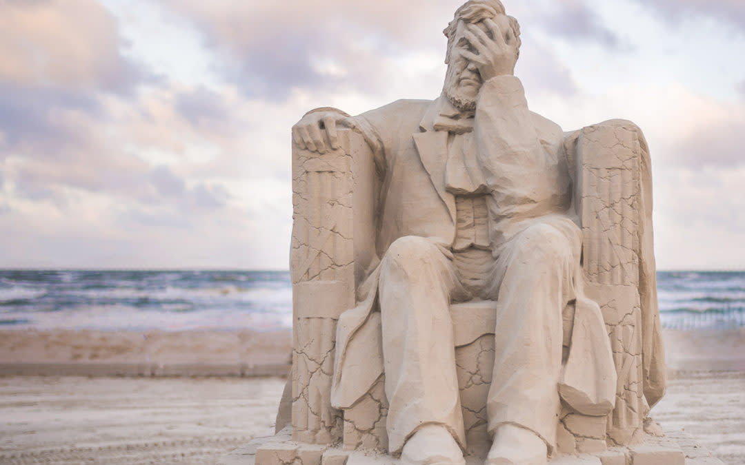 Horizontal photo of a sand sculpture on the beach depicting Abraham Lincoln sitting in a crumbling chair with his hand to his face