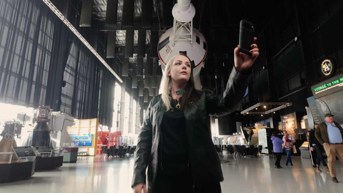 Phoenyx Powell taking a selfie with the Saturn V rocket at the US Space and Rocket Center in the background.