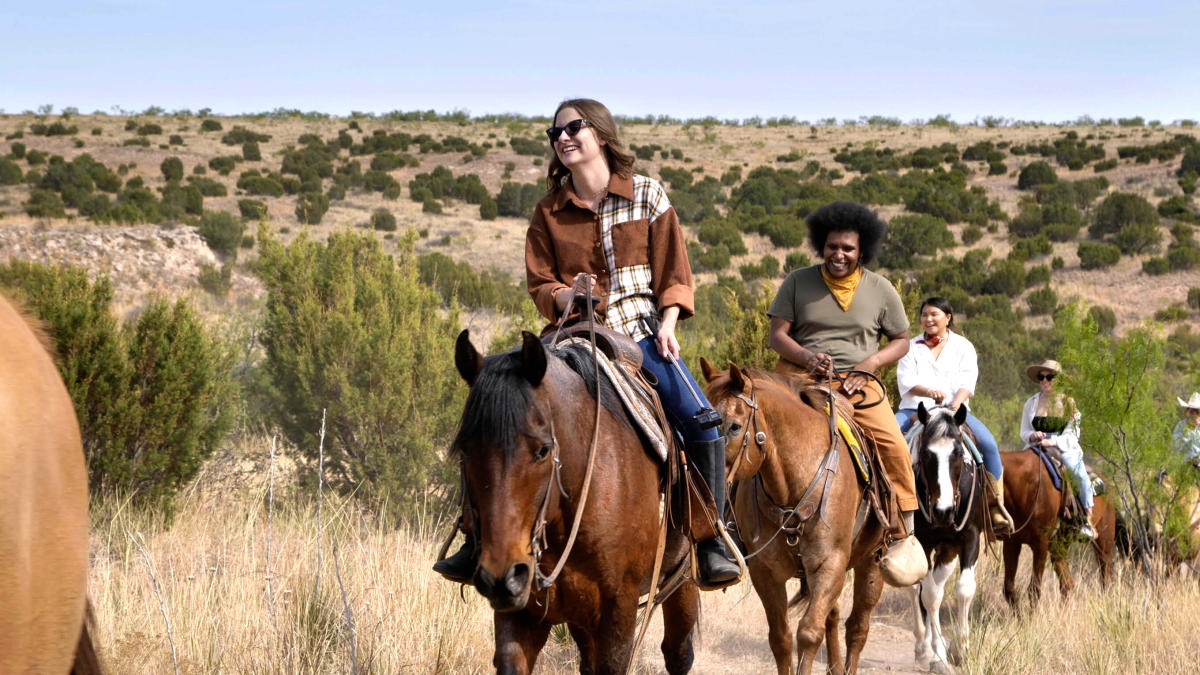 A woman with brown hair fair skin riding a dark brown horse with black hair, a man dark man riding a brown horse, and two women riding horses in the background at Los Cedros Ranch