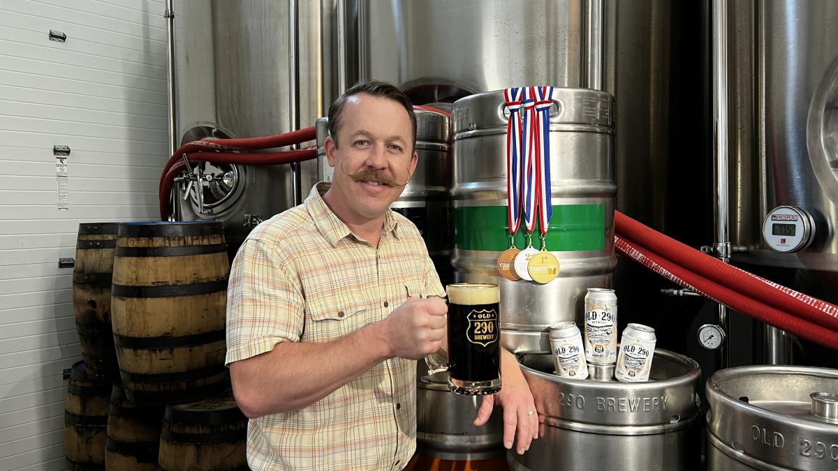 Image of a man holding an award winning beer in front of multiple kegs and winning medals.