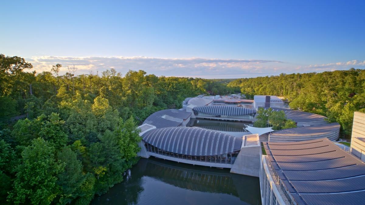 Aerial view of modern architectural structures nestled among lush greenery, reflecting on a serene waterway under a clear blue sky.