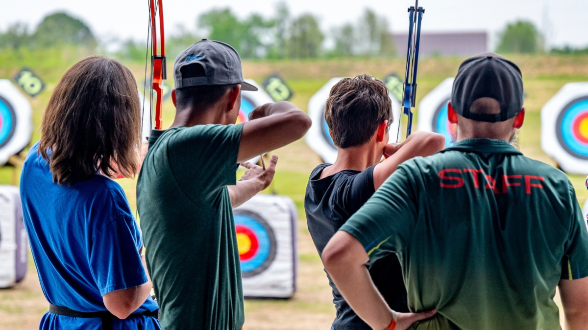 The image shows four individuals at The Quiver Archery Range in Osage Park, Bentonville, Arkansas, practicing archery and aiming at colorful targets.