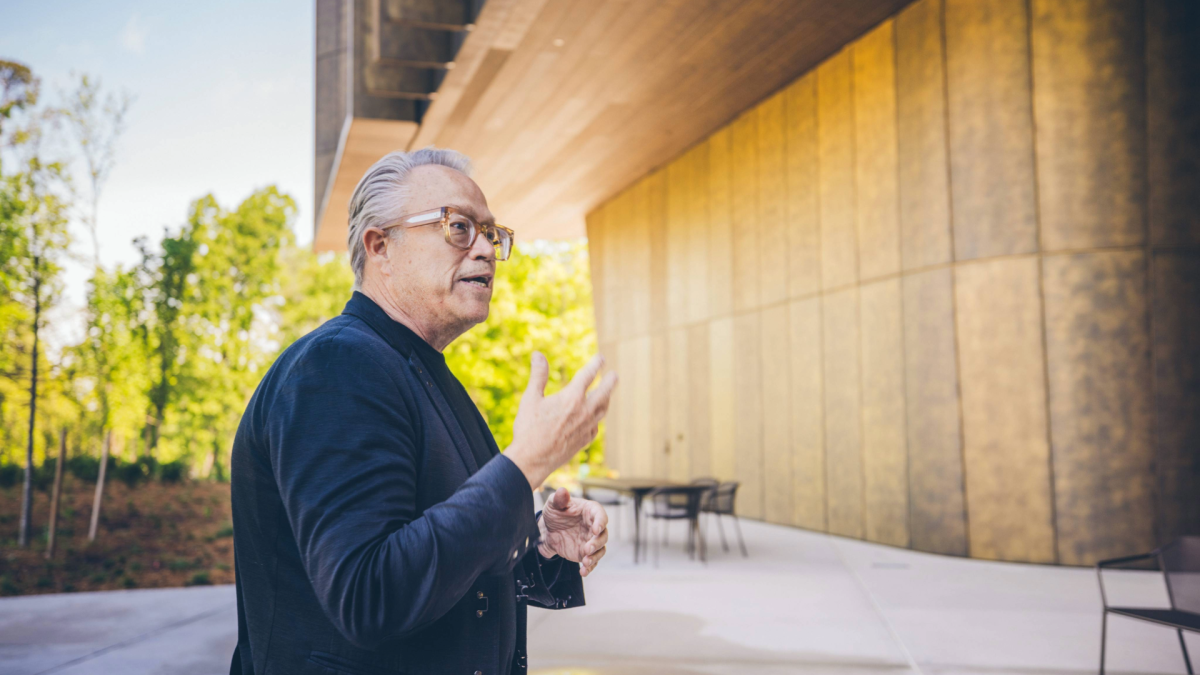 A man with gray hair and glasses gestures while speaking outside a modern building with curved golden walls. The scene is set on a sunny day with green trees in the background and patio chairs nearby.