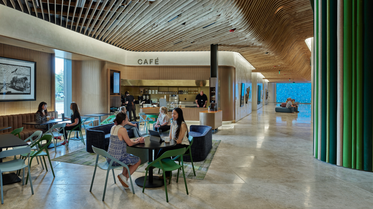 A contemporary cafe and lounge area with a curvilinear, slatted wood ceiling. People are seated at tables and in lounge chairs, some near a "CAFÉ" counter. Green and blue accents are visible, including a vibrant blue wall in the background.