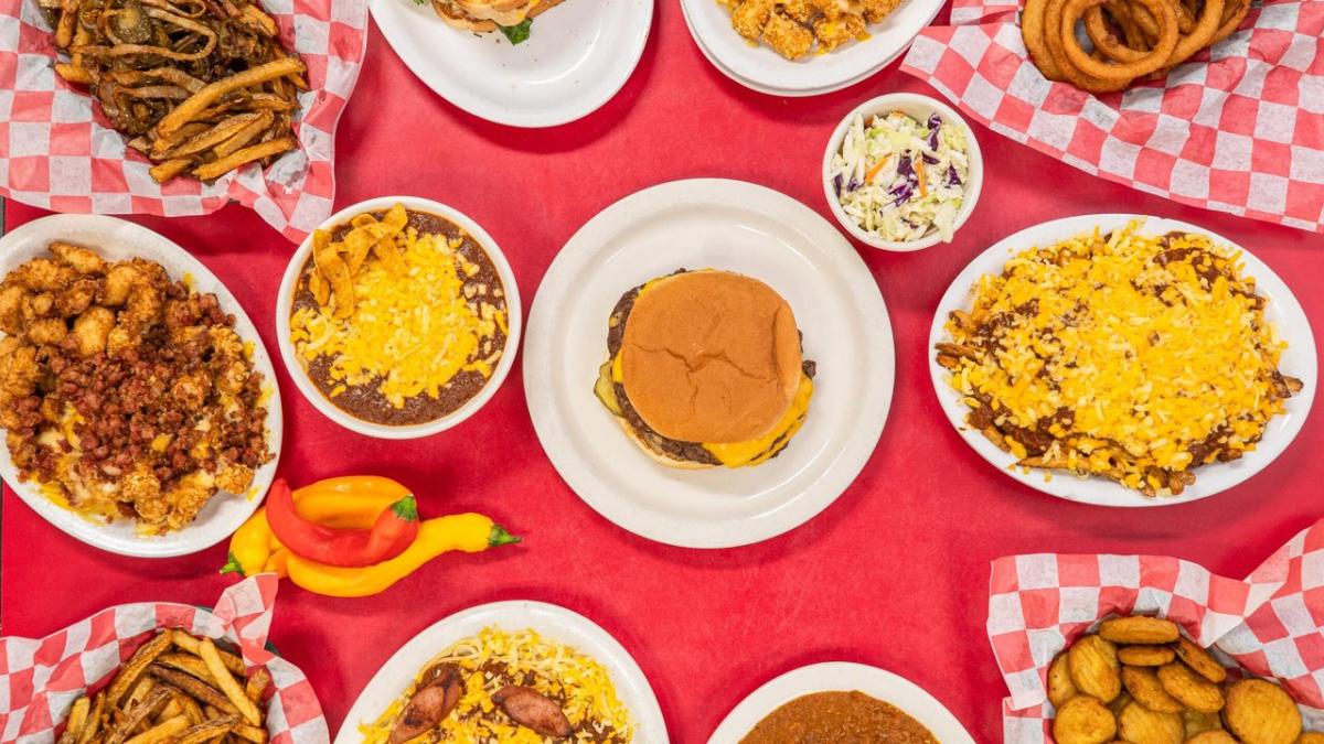 A vibrant spread of various dishes on a red table, featuring a cheeseburger, fries, and sides like coleslaw and chili.