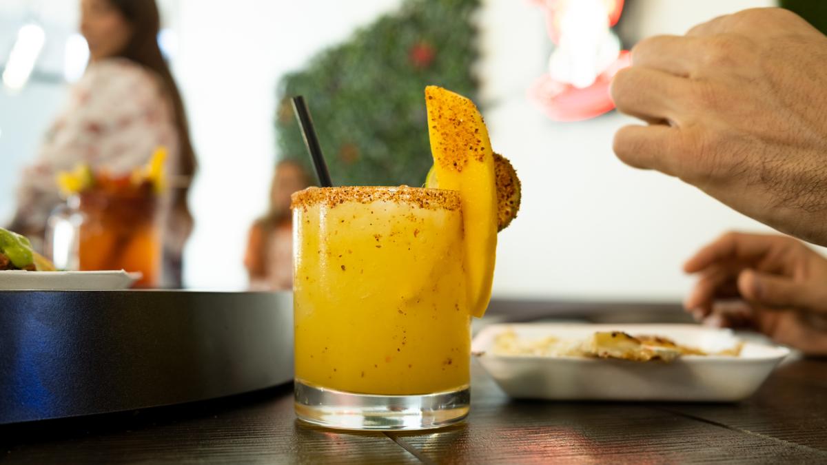 A yellow margarita in a short glass, garnished with a mango slice and tajin on the rip, sitting on a table with dinners eating from plates blurred in the background.