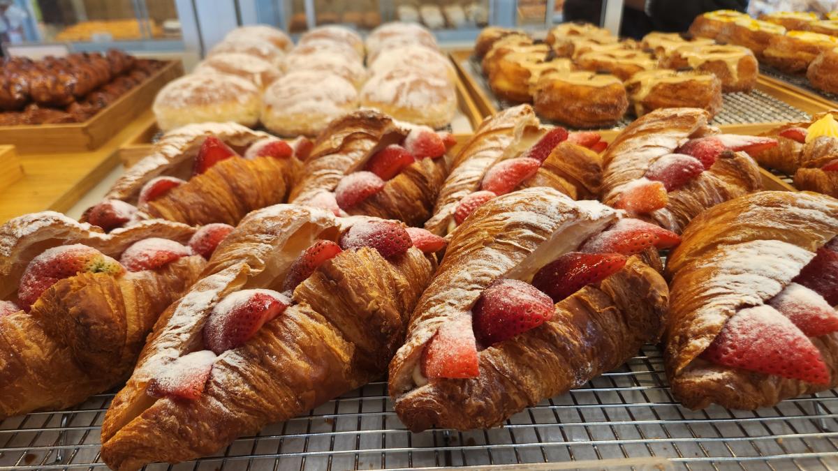 A bakery display with six golden croissants with a filling of custard and fresh sliced strawberries and dusted with powdered sugar in the foreground