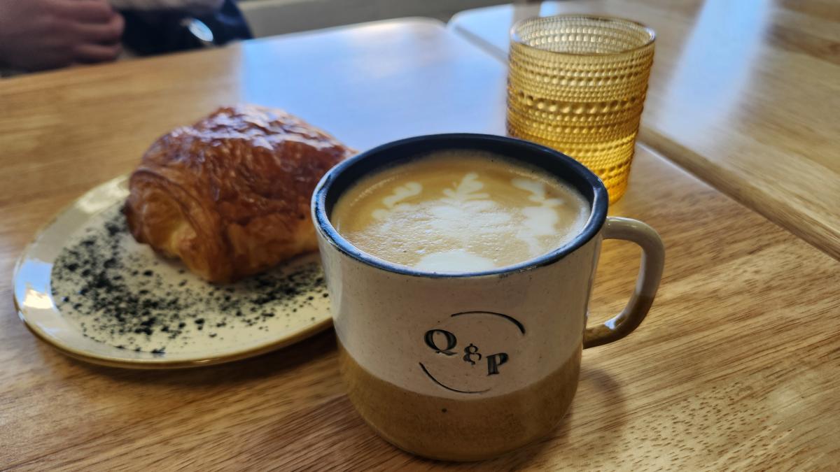 a Quiches and Pies branded coffee cup with a hot latte and a plated chocolate croissant and glass of water