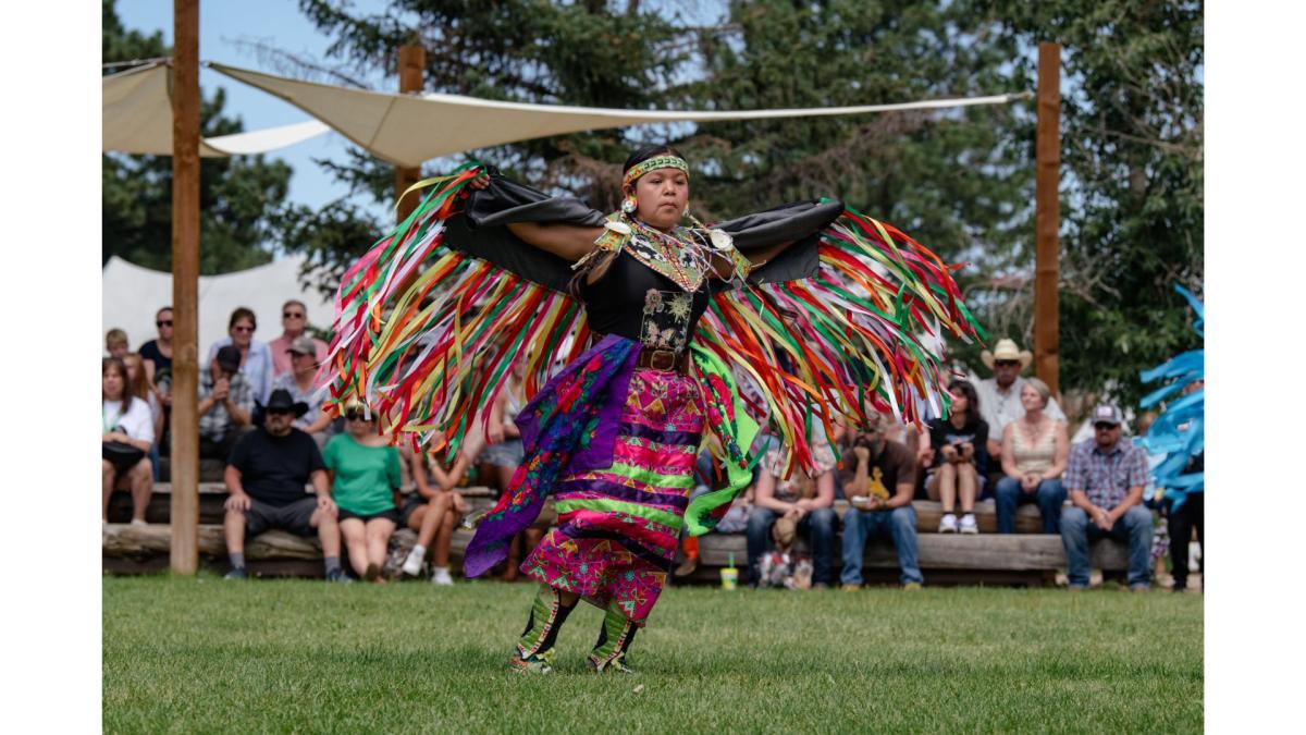 Native woman in vibrant shawl regalia performing a Fancy Shawl Dance with colorful ribbons at Morning Star American Indian Village.