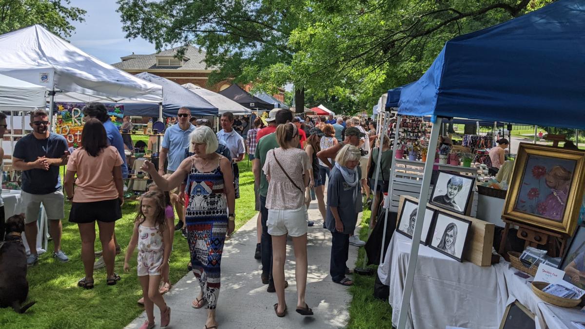 Image is of vendor tents set up on a sunny day with people walking by and shopping.