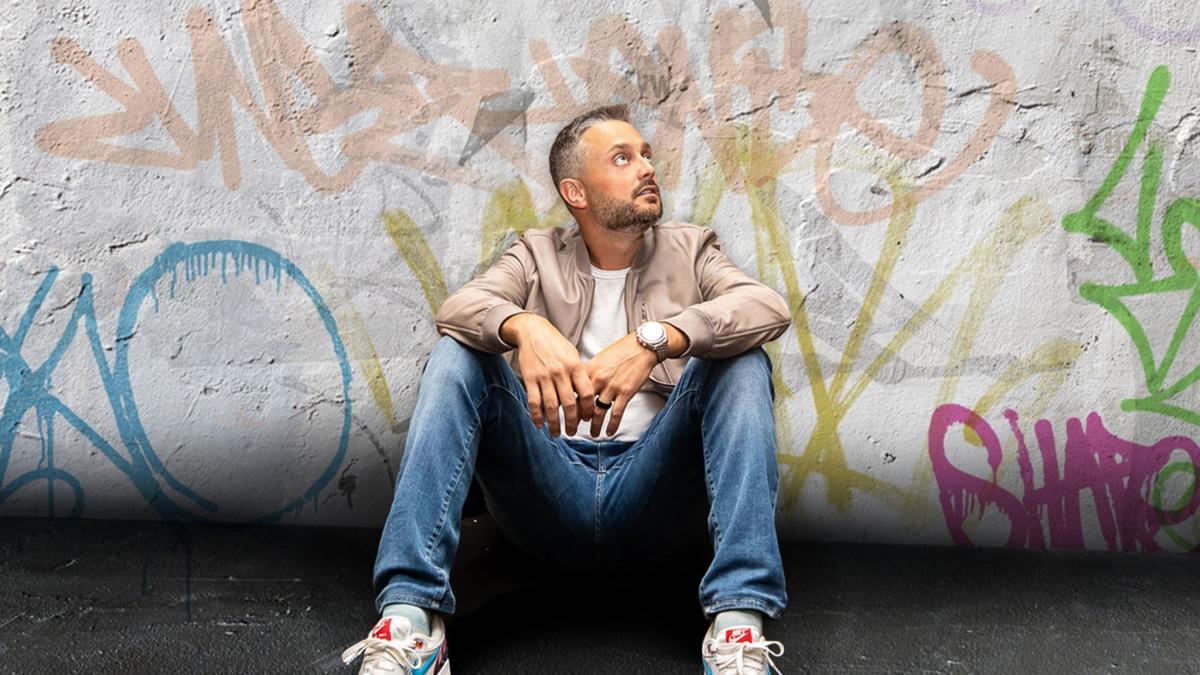 Image is of Nate Bargatze sitting up against a wall that is covered in graffiti.