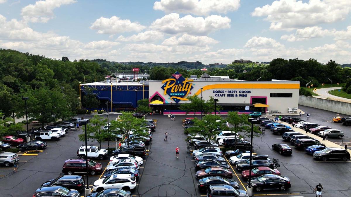 Image is of the front view and parking lot of the Party Source with cars in the lot and people walking during the day.