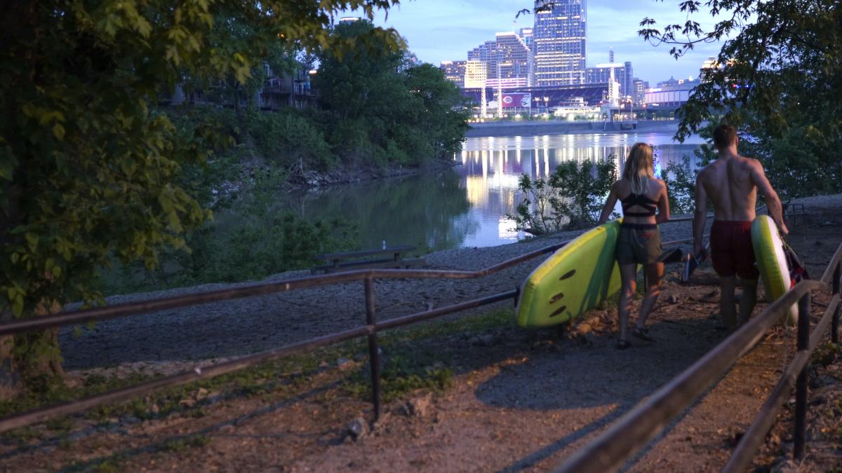 Image is of a couple carrying kayaks with the Licking River to the left and the City of Cincinnati in the foreground.
