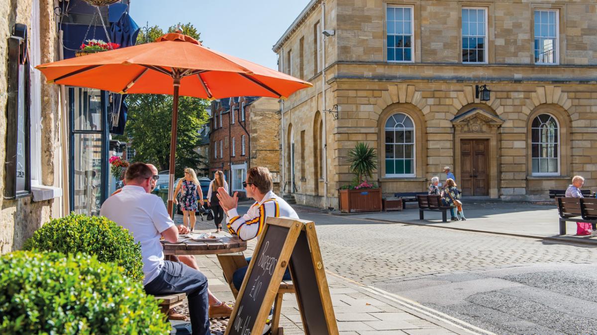 A group of people sit in the shade of an orange parasol outside a cafe, people sit on benches outside the adjacent town hall, whilst two ladies walk along shopping. Woodstock in the Cotswolds