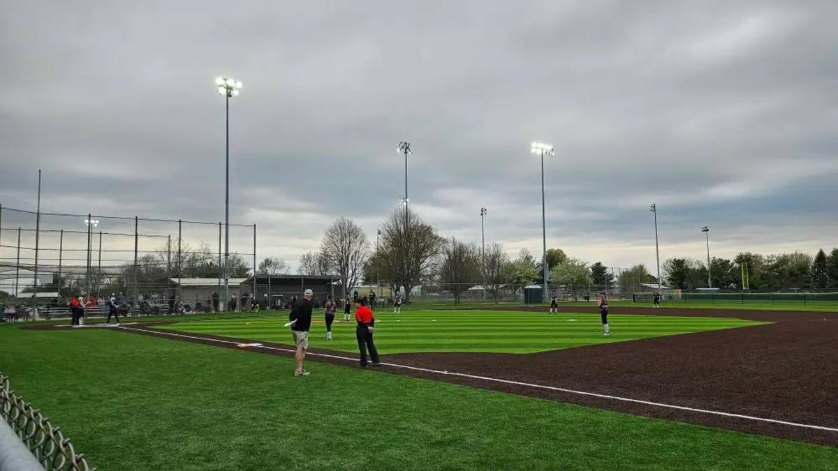 Youth softball game on a turf field under stadium lights with overcast sky.