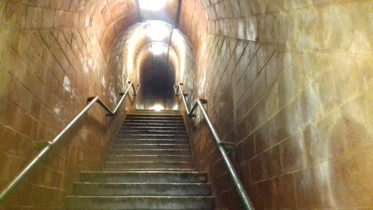 An underground tunnel with a narrow staircase and metal handrails, leading upwards. The dim lighting creates a mysterious and eerie atmosphere.