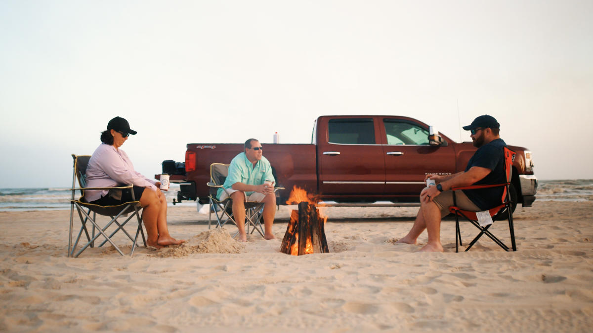three adults sitting on the beach with a bonfire