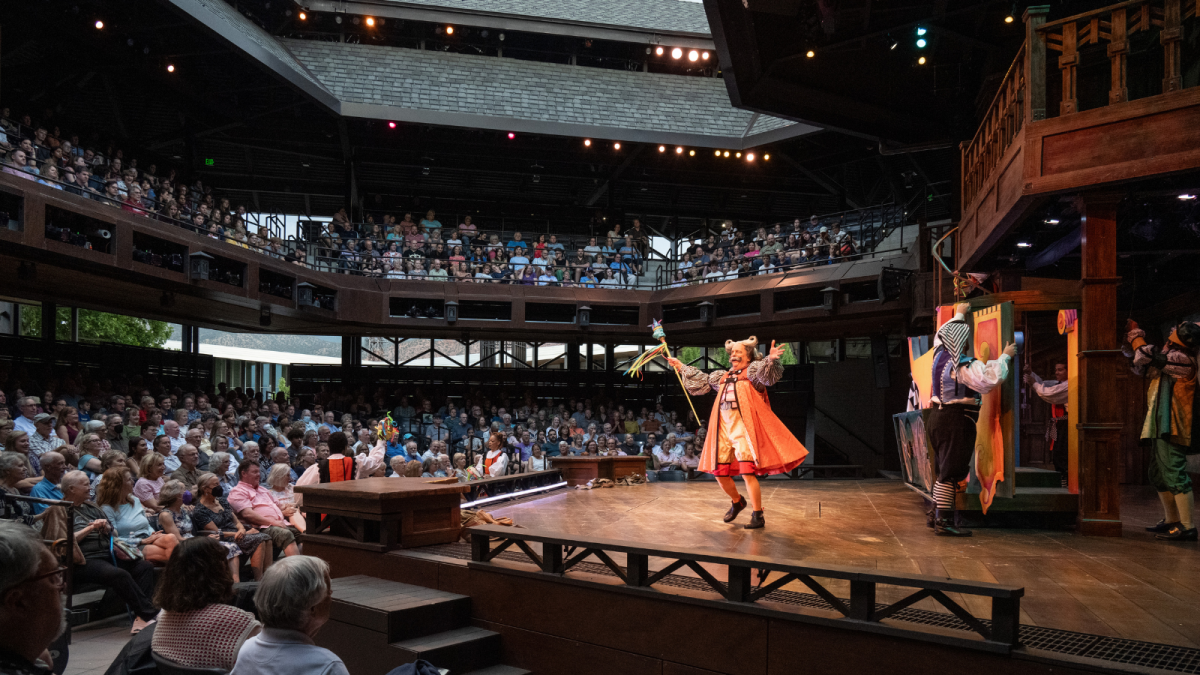 A crowd sits in an Elizabethan-style outdoor theater around a large wooden stage with actors dressed in vibrant Shakespearean garb, arms wide in mid-performance.