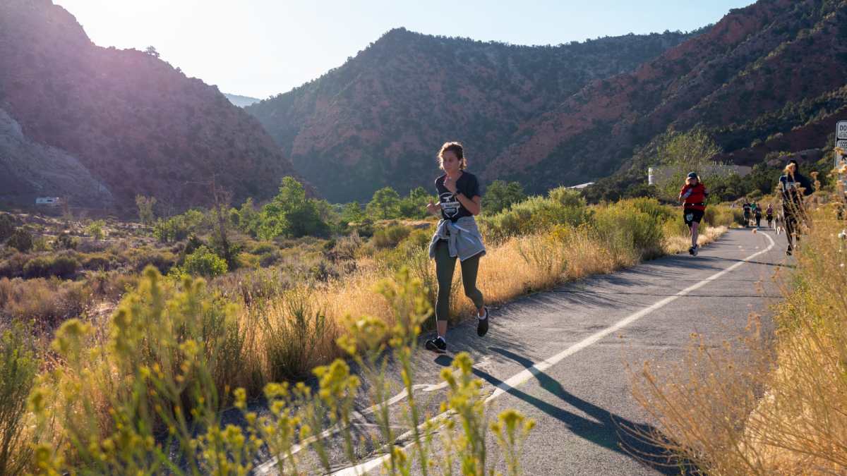 A handful of marathon runners follow a paved trail down Cedar Canyon for the Cedar City 1/2 Marathon with the sun rising behind them and the red rock views of the canyon visible behind them.