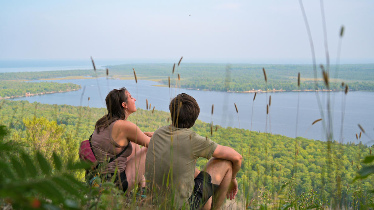 two people side trailside and look out over the forest and lake below