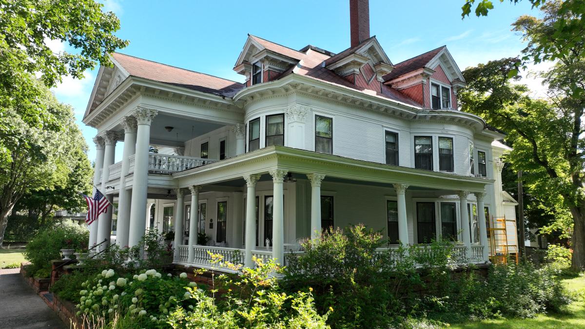 historic mansion with wrap around porch and columns is framed with lush summer foliage and flowers