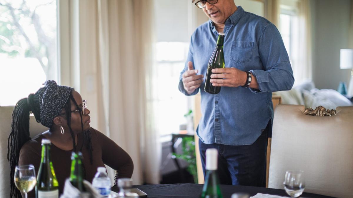Person holding a bottle of wine talking to individual sitting at a table during a wine tasting