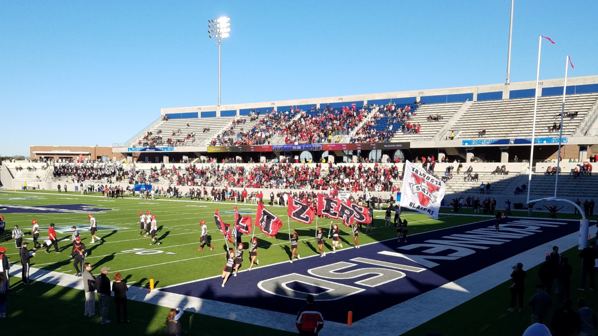 Football players on a turf field in a stadium with cheerleaders running with flags across the field