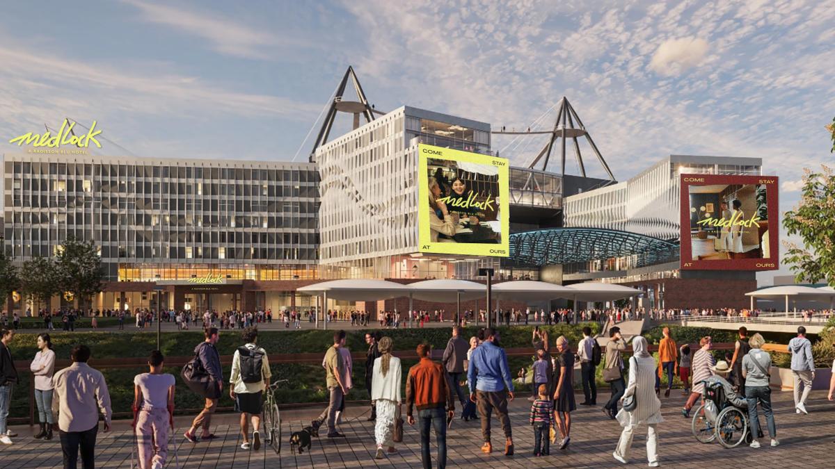 Modern hotel complex beside a large stadium with distinctive triangular roof structures, crowds gathering in a landscaped plaza. Branding for “Medlock, a Radisson Blu Hotel” appears on the building, with colourful promotional posters displayed on the exterior.