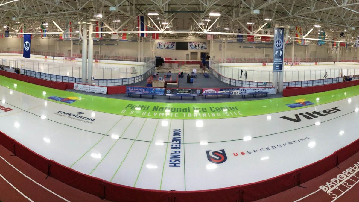 The interior of the Pettit National Ice Center with white ice adorned with decals and lines in the foreground, two ice rinks in the midground and a long tan wall in the background and lights shining down reflecting off the ice throughout.